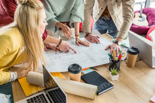 Preview: group of female and male architects working on blueprint at desk with coffee to go in loft office