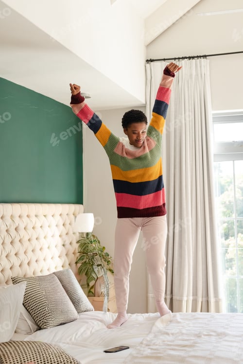 Preview: African American woman jumping on bed in striped sweater and pink pants, smartphone on bedding