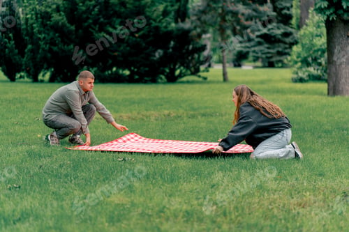 Preview: In the park a guy and a girl in warm clothes lay out a checkered blanket on the green grass seasonal