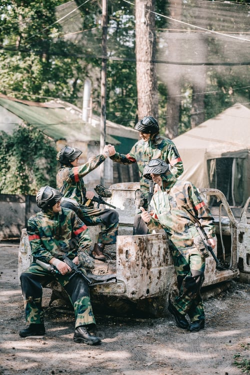 Preview: paintball team in camouflage and goggle masks celebrating victory and shaking hands on broken car