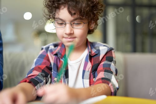 Preview: Portrait of concentrated latin boy in glasses writing while sitting at the desk and doing homework