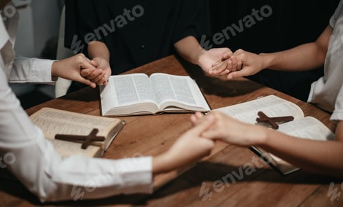 Preview: Group of Christians sit together and pray around a wooden table with blurred open Bible pages