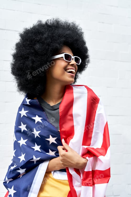 Preview: Happy young African woman wrapped in USA flag standing against white wall.