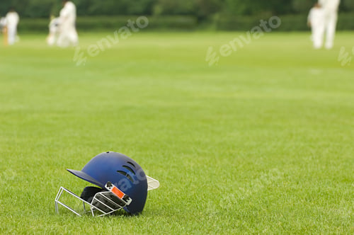 Preview: A cricket helmet rests on a lush green field with blurred players in the background.