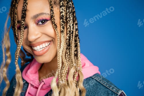 Preview: attractive african american woman in pink hoodie and denim vest looking at camera, blue background
