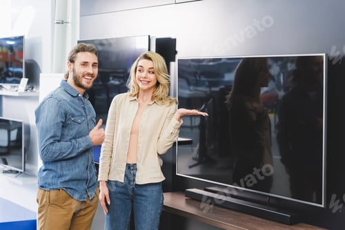 Preview: smiling boyfriend showing like and girlfriend pointing with hand at tv in home appliance store