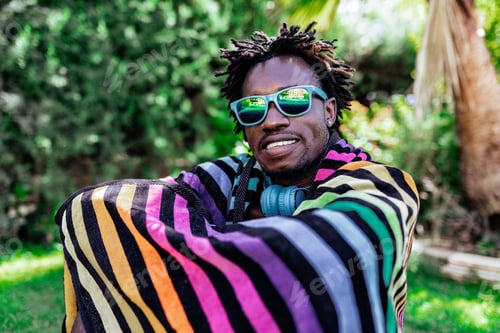 Preview: Smiling African American male in sunglasses wrapped in colorful towel sitting on meadow in summer