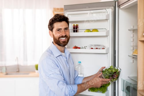 Preview: Glad hungry attractive adult caucasian man with beard takes out fresh salad from refrigerator, enjoy