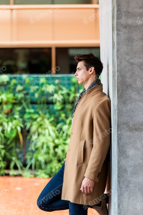 Preview: Side view of trendy young man wearing coat and scarf leaning on a wall while looking away