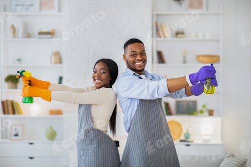 Preview: Portrait of young black couple using detergent spray bottles as guns, posing during cleanup at home