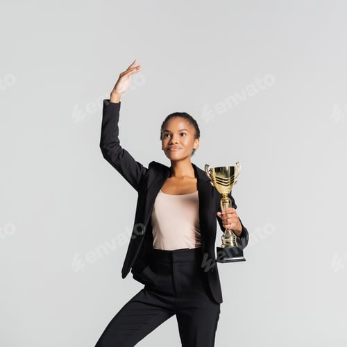 Preview: smiling african american businesswoman performing ballet with golden cup isolated on grey
