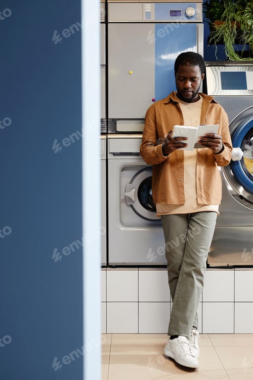 Preview: Young African American man in casualwear standing by washing machine