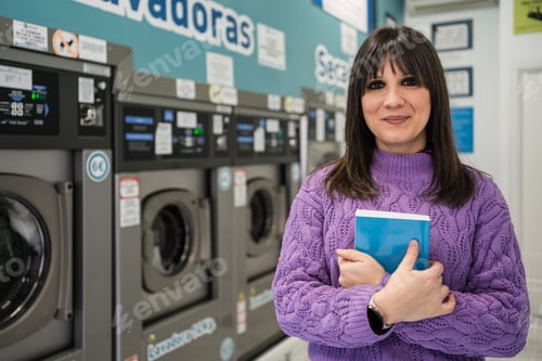 Preview: Young woman holding a book waiting for laundry in laundromat