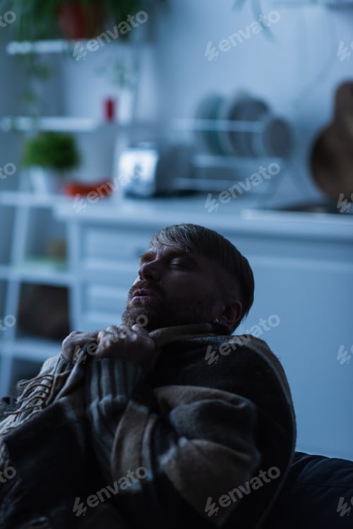 Preview: frozen man wrapped in warm blanket sitting in blurred kitchen with closed eyes