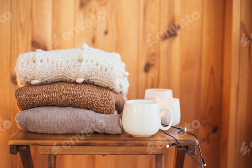 Preview: Stack of folded cozy knitted sweaters on a table near mug with hot tea and candles in a wooden house