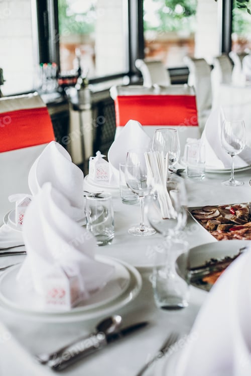 Preview: Wedding table with food, silver cutlery, empty glasses, white silk chairs and napkins