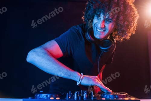 Preview: Man with curly hair using DJ equipment and standing in the dark neon lighted room