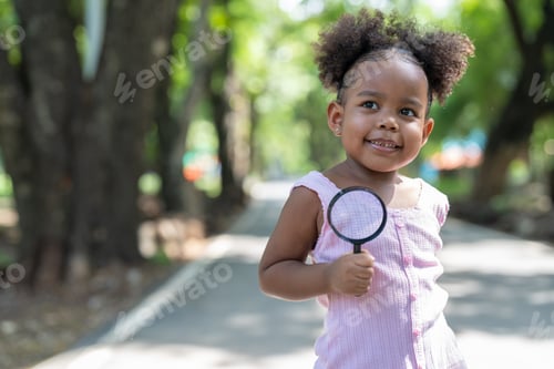 Preview: Smiling African American little girl holding magnifying glass to explore on the tree in park