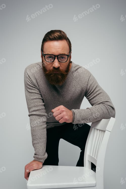 Preview: Bearded man in eyeglasses looking at camera while posing near chair isolated on grey
