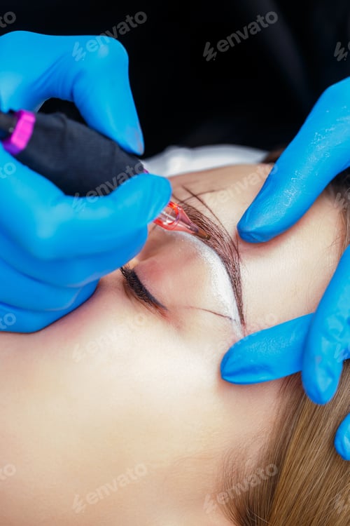Preview: Beautician tattooing a woman's eyebrows using special equipment during permanent make-up, close-up.