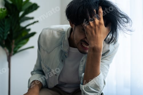 Preview: Asian young upset depressed man sitting alone in living room at home.