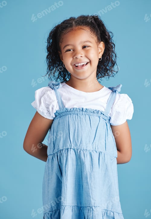 Preview: Happy smiling little girl standing with her hands behind her back against blue studio background. C