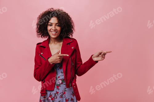 Preview: Young girl in red jacket and flowered dress shows fingers on place for text on pink background. Por