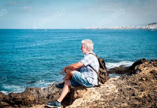 Preview: Side view of attractive senior man casual dressed sitting in front to the sea. White hair and beard.