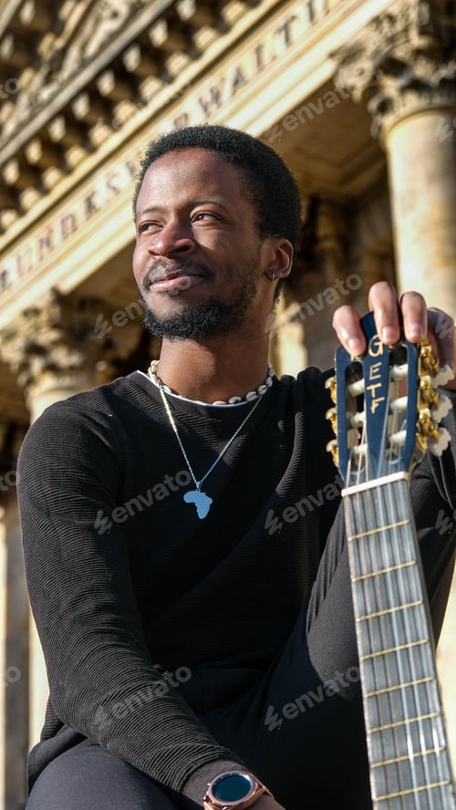 Preview: Young African man holding a guitar posing on a street in front of a stone building