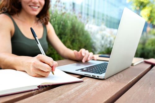 Preview: Close up of female college student writing on notebook with pen. Young woman using laptop outdoors.