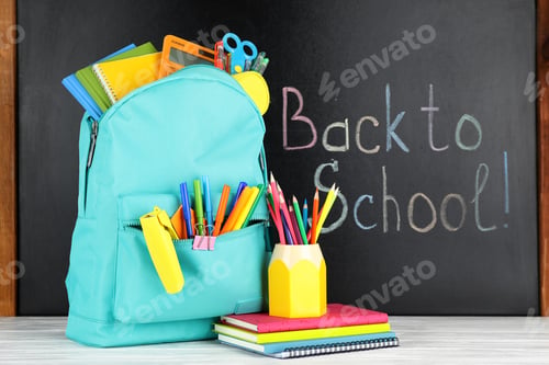 Preview: Bright backpack with school stationery on white wooden table near black chalkboard. Back to School