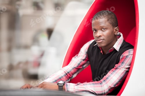 Preview: Young handsome african man in stylish casual clothing sitting in big red egg shaped armchair