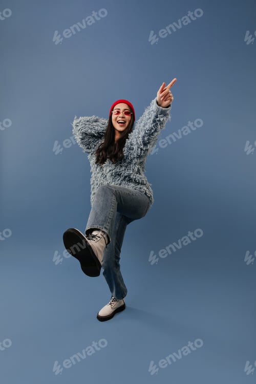 Preview: Positive long dark haired lady with red hat and glasses in trendy sweater, blue jeans and light coo