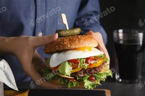 Preview: Woman holding tasty burger with fried egg over table, closeup