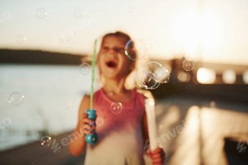 Preview: Amazing sunlight. Happy little girl playing with bubbles near the lake at park