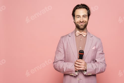 Preview: Young host of event holding microphone and looking at camera isolated on pink