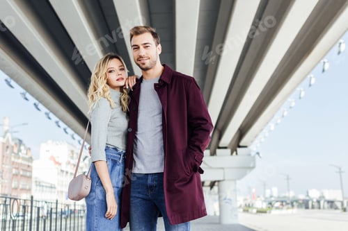 Preview: stylish couple in autumn outfit hugging under bridge in city and looking at camera