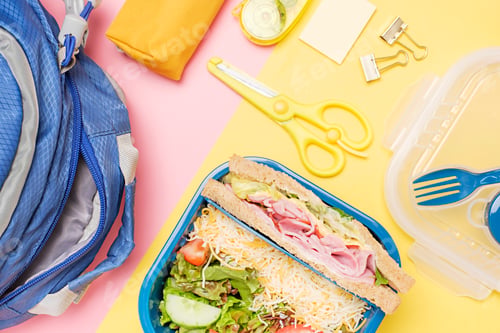 Preview: Backpack and plastic lunchbox with sandwich and salad on pink and yellow background top view.
