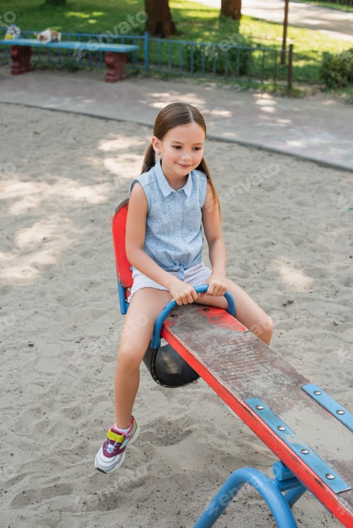 Preview: smiling girl in shorts riding seesaw in summer park