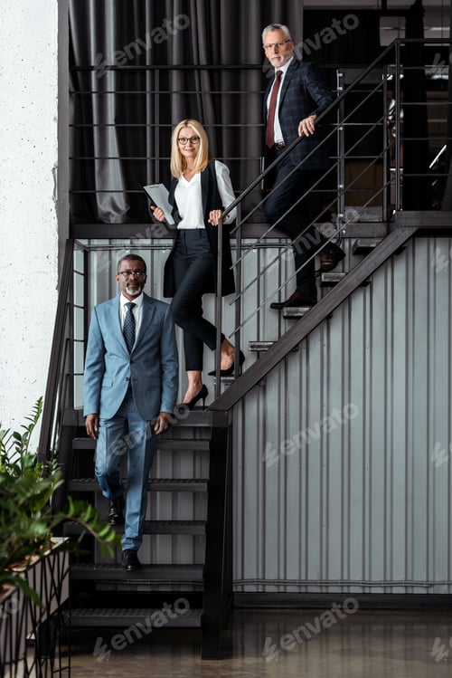 Vista previa: alegre empresaria rubia caminando por las escaleras cerca de empresarios multiculturales