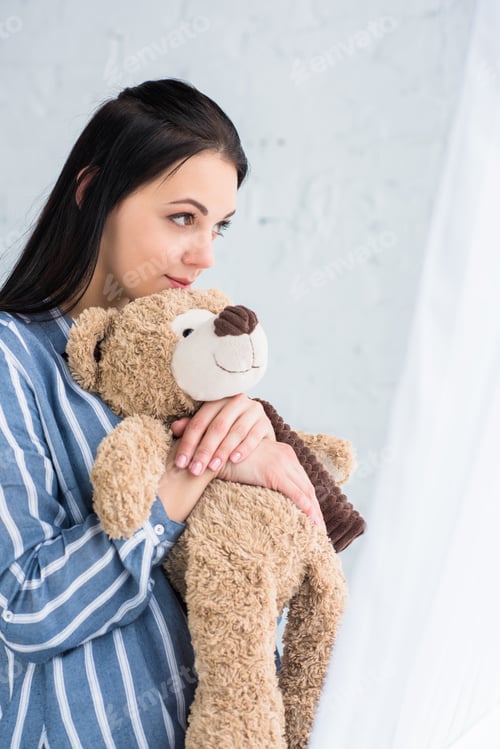 Preview: side view of pensive woman with teddy bear in hands looking away at home