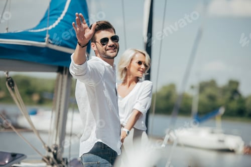 Preview: happy young man in sunglasses smiling at camera and waving hand while holding hands with beautiful