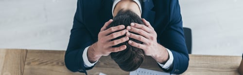 Preview: panoramic shot of depressed businessman holding hands on bowed head while sitting at desk in office