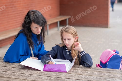 Preview: smart girl solving math problems on her own while her mother sits nearby at a wooden table on