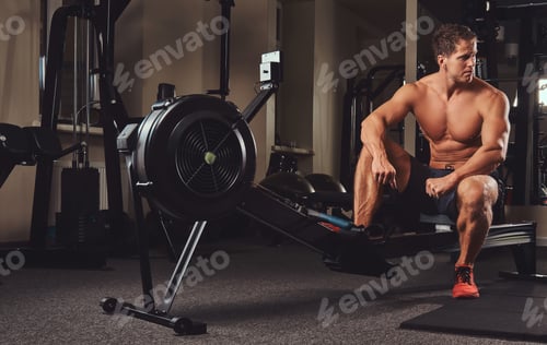 Preview: Muscular shirtless athlete resting after a hard workout while sits on the rowing machine in the gym.