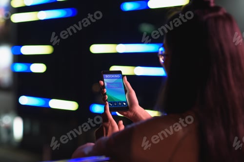 Preview: back view of girl holding smartphone with booking appliance on street with neon light in evening,