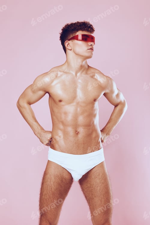Preview: Confident young man posing in stylish sunglasses and white swimwear against a pink background