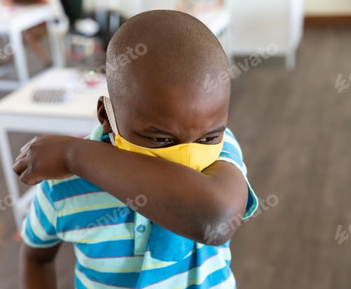 Preview: African American boy coughing into elbow, wearing yellow face mask in classroom with white tables