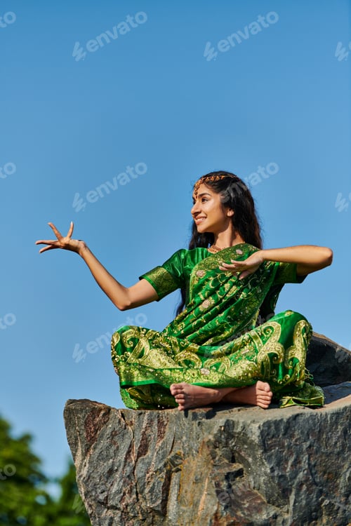 Preview: cheerful and stylish indian woman in green sari posing on stone with blue sky on background