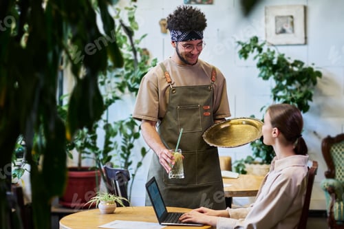 Preview: Young waiter with glass of refreshing lemonade looking at pretty female guest